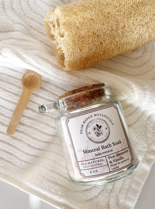 Jar of Pear Ridge Botanicals Mineral Bath Soak with a loofah and wooden spoon on a textured surface